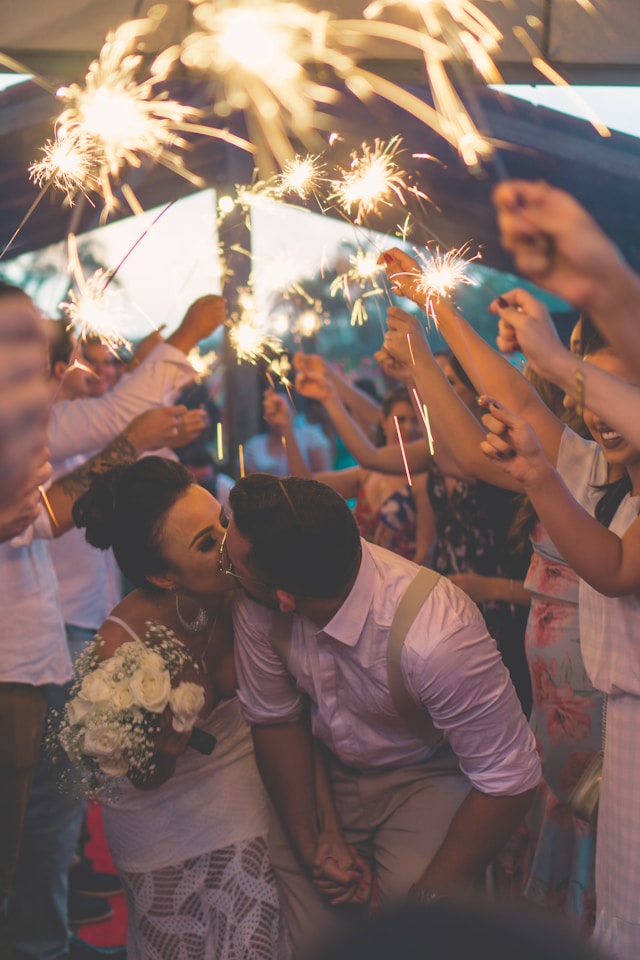 Novomanželé při polibku na svatbě pod zapálenými prskavkami, které drží jejich přátelé a hosté. Zdroj: unsplash.com